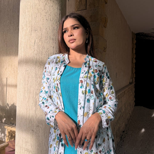 Woman wearing a floral jacket over a blue top, standing against a stone wall.