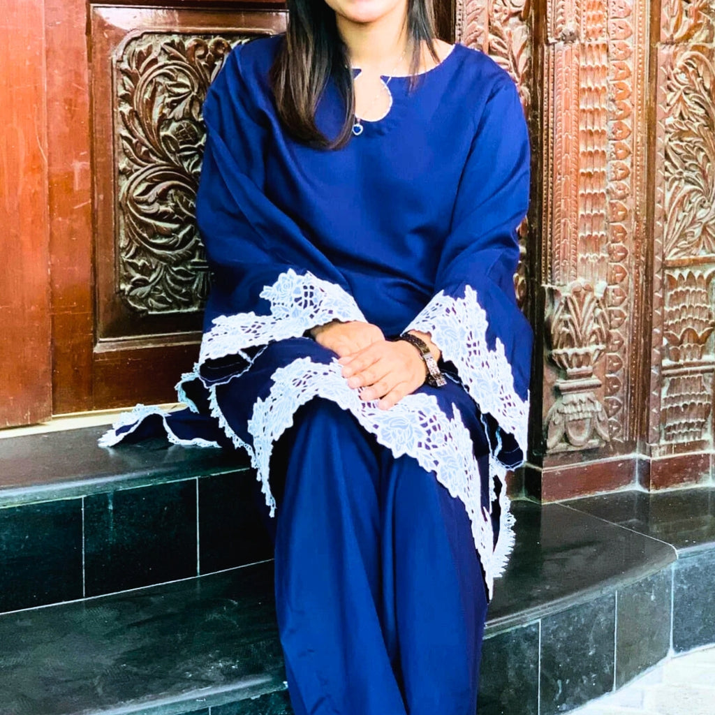 Woman in a blue dress with lace sleeves sitting against a decorative wooden wall.