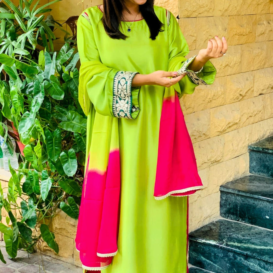 Person wearing a bright green and pink traditional outfit standing outdoors with plants and steps in the background.