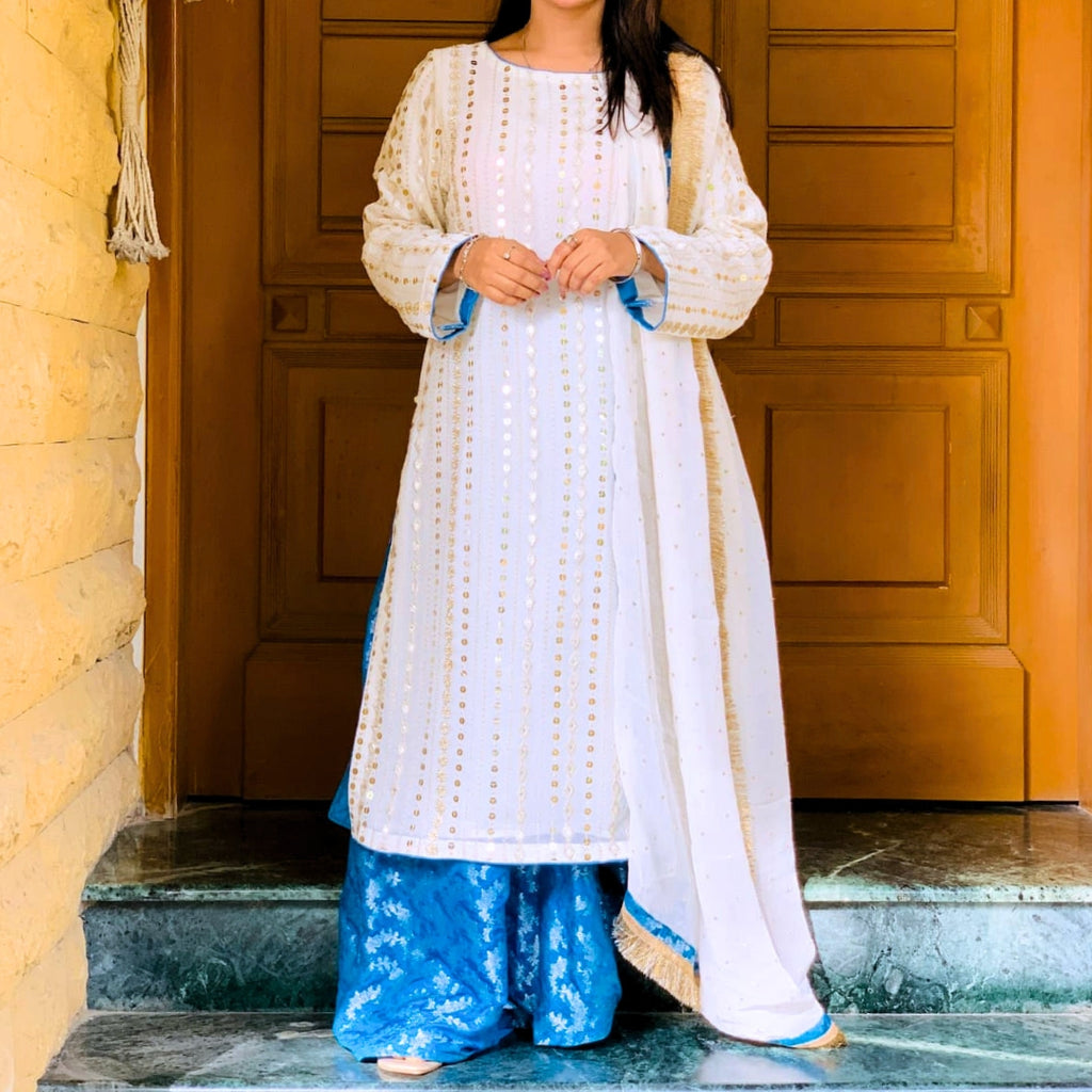 Woman wearing a white and blue traditional outfit standing in front of a wooden door.