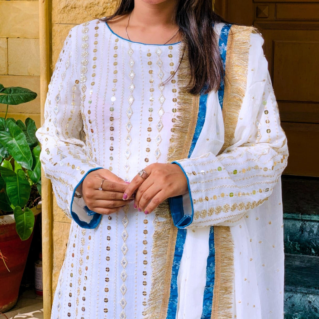 Person wearing a white and gold embroidered outfit with blue accents, standing indoors.